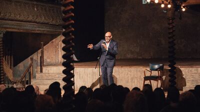 A performer wearing a suit stands on a wooden stage speaking into a microphone to a large audience