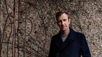 Robert Macfarlane stands against stone background wearing a navy blue coat