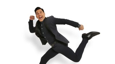 Russell Kane wears a black suit against a white background.