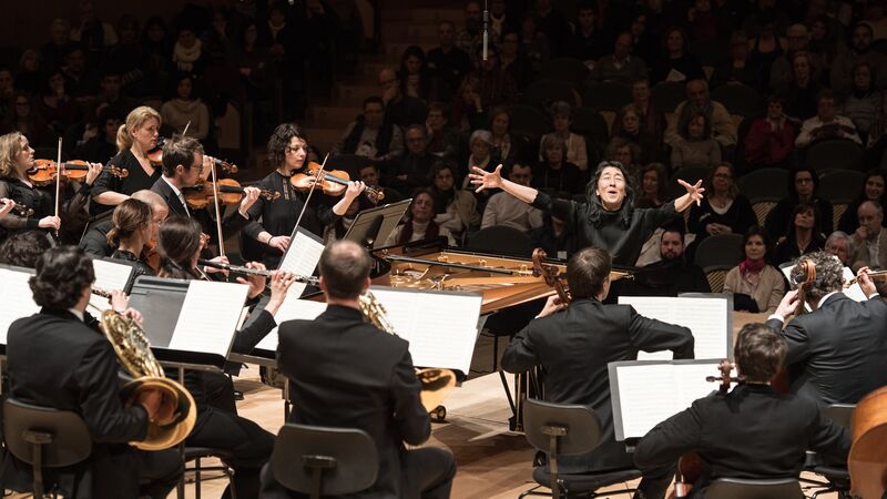 Mitsuko Uchida with open arms facing the Mahler Chamber Orchestra. Audience are sitting behind her in the shot