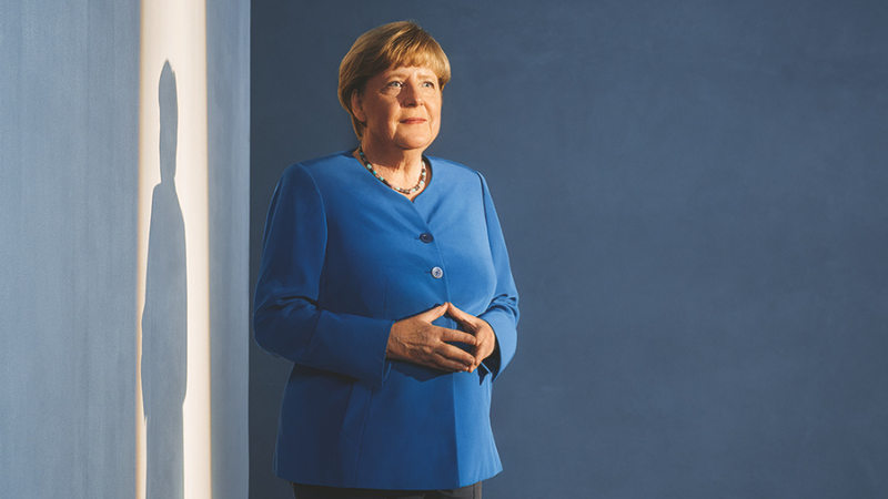 Angela Merkel standing by a wall in a blue jacket.