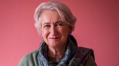 Lindsey Hilsum stands against a pink background wearing a green jumper and scarf.