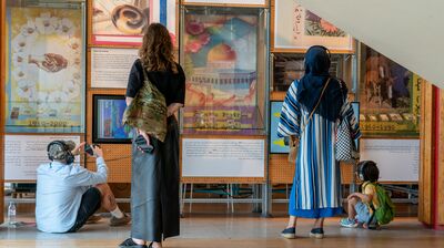 Visitors explore a cultural exhibit with vibrant posters, artifacts, and Arabic text. A child and adults listen intently with headphones