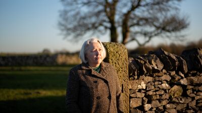 Woman in a brown jacket stands by a wall in a field.