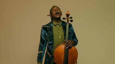 A man holds a cello neck in his right hand and his cello bow in his left hand. His eyes are shut and he is looking up. His mouth is open as though he's singing.