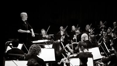 A conductor standing in front of an orchestra seated behind music stands playing violins