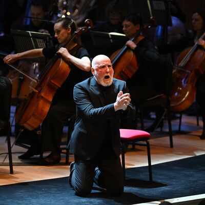 An singer wearing a suit on his knees on stage with his hands clasped together. An orchestra is playing behind him.