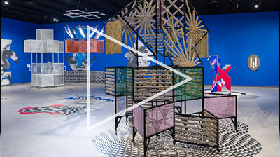 Installation view of colourful wicker sculptures on wheels, in a large room with a blue wall including black and white cut out images behind