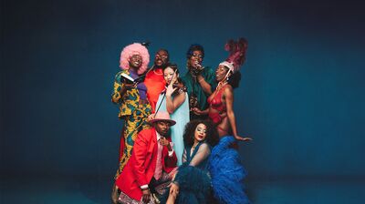 Group of 7 performers wearing colourful and feathered outfits posing for a photo against a blue background