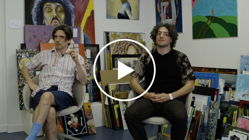 Artists Jeremy Deller and John Costi sit on chairs in the Koestler Arts building, around them multiple artworks (predominantly paintings) adorn the walls; others are propped up in stacks against the base of the walls.