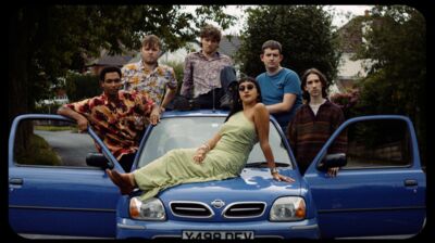 Group of people in colourful clothing surrounding and sitting on a blue Nissan car