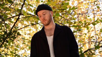 Artist wearing a black cap and blazer gazing down, with a background of autumnal leaves