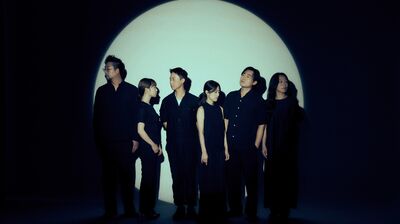 Six people wearing all black stand grouped together in a circle of light in an otherwise dark room