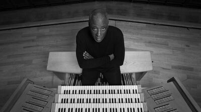 An organist sat behind an organ with their arms crossed looking up at the camera