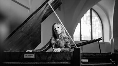 A pianist in a red leather jacket leaning behind a grand piano inside a wite room with a large window.