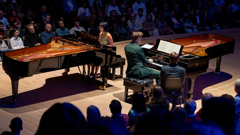 Víkingur Ólafsson & Yuja Wang each sit at a iano. The pianos are facing different directions, while they are almost next to each other. Audience members are on all sides of the Royal festival Hall stage.