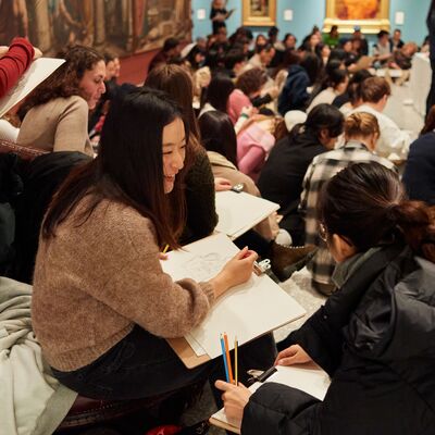 A room of people sitting, drawing. They are holding clipboards and pencils. One person looks at their neighbour and smiles.