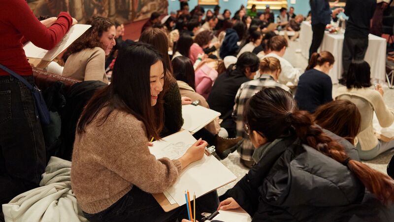 A room of people sitting, drawing. They are holding clipboards and pencils. One person looks at their neighbour and smiles.