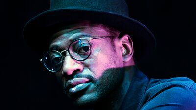 Artist Mark Kavuma wears a hat and black rimmed glasses as he poses for a photo in a dark room