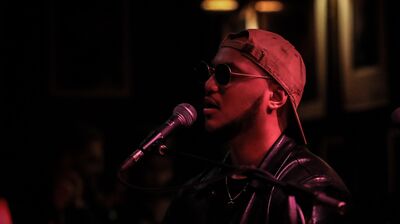 A man wearing a baseball cap and sunglasses singing into a mic playing keyboard