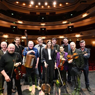 A group of musicians holding various instruments (violins, accordions, percussion) on a stage