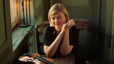 A young woman with blonde hair sits at a wooden table, smiling softly, with a violin and bow resting beside her