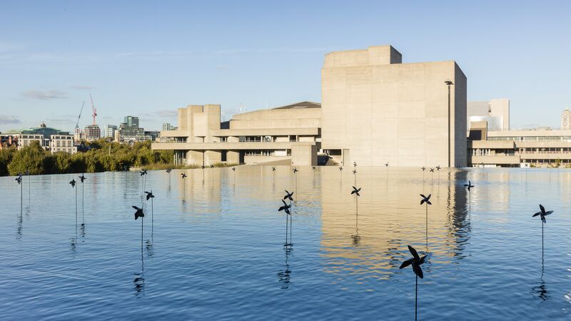 Installation view of small paper windmill toys suspended in water with a view of concrete buildings behind