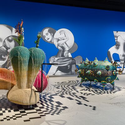 Installation view of colourful wicker sculptures on wheels, in a large room with a blue wall including black and white cut out images behind