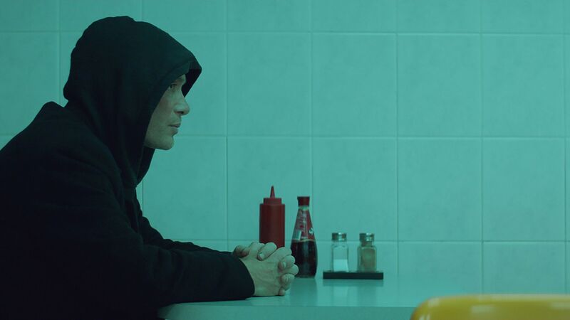 A male actor sat at diner table, wearing a black jacket and black hoodie