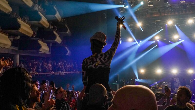 Grace Jones crowd-surfs in a packed Royal Festival Hall auditorium. She if lifted high and raising her hand in the
air holding a microphone. You can see the back of her head wearing a sparkly bowler hat. Lights shine down and she has confetti over her.