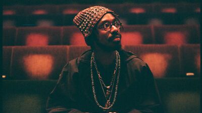 Artist SHABAKA sits in an empty auditorium holding a wooden flute in his hands as he gazes off camera.