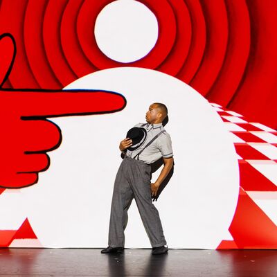 a performer in grey trousers and shirt with braces on stage leaning away from a large red hand stage prop