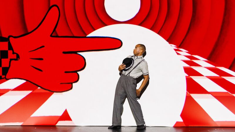 a performer in grey trousers and shirt with braces on stage leaning away from a large red hand stage prop