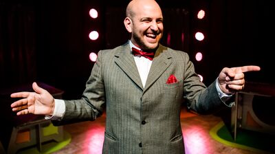 Nick Cassenbaum is centred in the image, dressed in a suit. He's standing on stage, laughing warmly and pointing to the right.