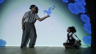 a performer in grey trousers and shirt with braces on stage holding out a hand for blue cartoon bird