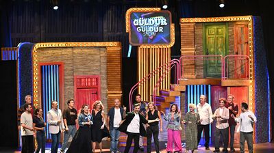 Large group cast on stage all looking toward one person in the middle, a sign for Gulder Gulder show hangs above