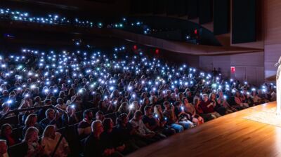 Performer wearing a top with white hanging sleeves, taking a selfie in front of an audience with their camera lights on