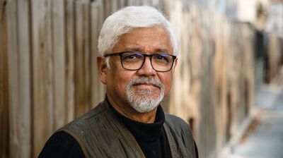 A person with white hair and glasses with facial hair wearing a gilet standing by a wooden fence.