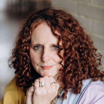 A person with red curly hair and blue eyes wears a multicoloured top with her head on her hands.