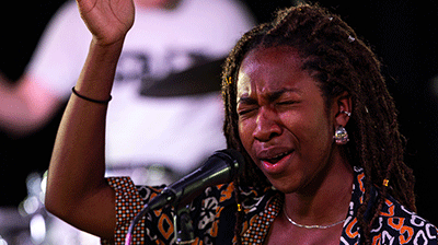 A young vocalist in a colourful top singing with eyes closed and one arm raised