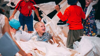 Children smile and laugh, moving through a space filled with torn up paper across the floor.