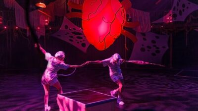 Two performers dressed in white costumes and white hats are holding hands and moving around a box, with inflatable hanging heart and hanging material suspended above them, lit by purple and blue stage lights.