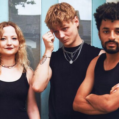 Three people each wearing a black t-shirt posing in front of a blue door. One has long blond hair, one with short blond hair and another with dark hair