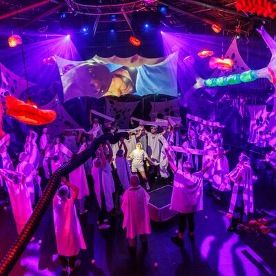 A group of performers on stage, holding white banners in the air with floating inflatable organs hanging from the ceiling