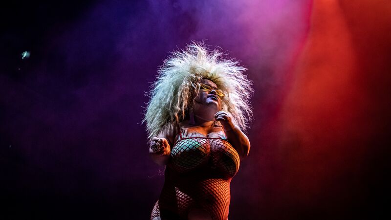 Midgitte Bardot stands in a fishnet dress and green bra with bright blonde hair that fans out against the purple and red lit backdrop.