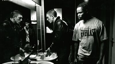 Black and white film still of three people in a bathroom, one washing their hands looking in the mirror