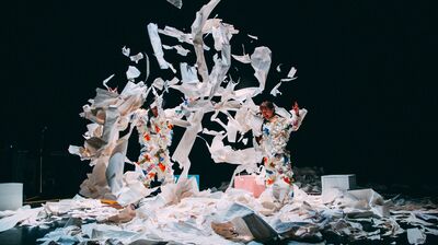 Two performers throw piles of paper up in the air and all around them. They are in costumes that are made of paper.