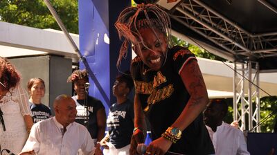 A woman dancing on stage outside, with a man playing an instrument and backup singers in the background