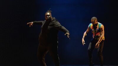 Two people dancing on a dark stage. One wears a black suit and the other wears a colourful waistcoat.