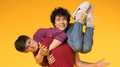 One performer in a red t-shirt smiles at the camera as they carry another performer in a purple t-shirt on their shoulders.